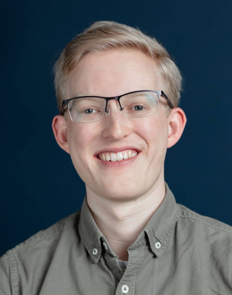 A person with short blond hair and glasses smiles at the camera, wearing a gray button-up shirt against a dark blue background.